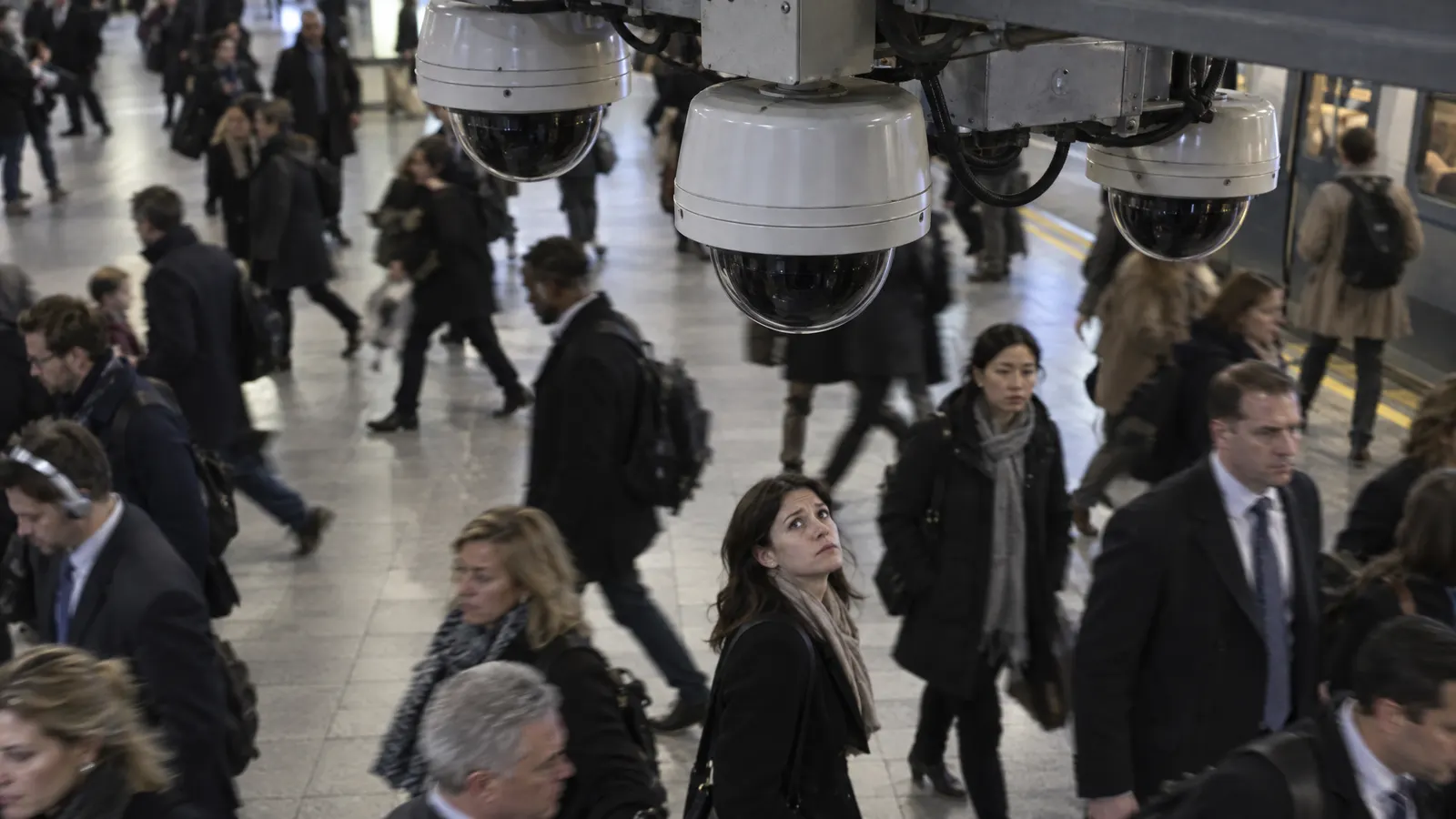 Surveillance cameras above a crowded train station while a woman looks up at them — illustrating AI facial recognition and mass surveillance