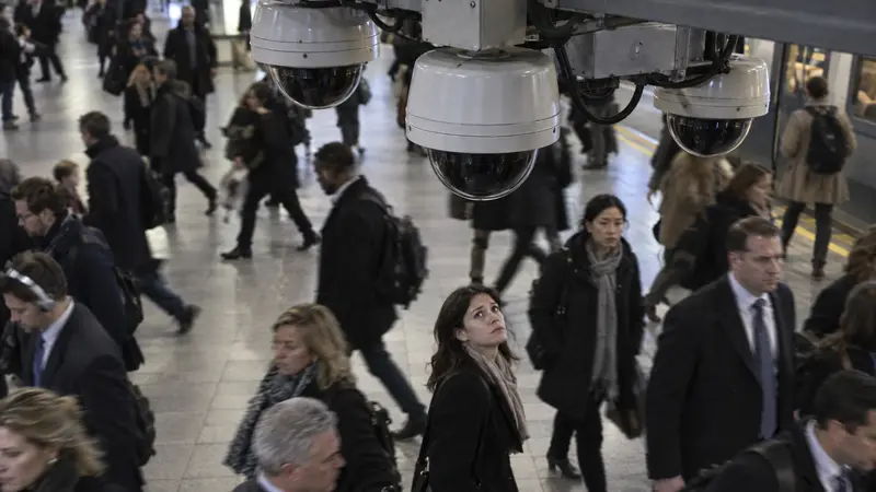 Surveillance cameras above a crowded train station while a woman looks up at them — illustrating AI facial recognition and mass surveillance